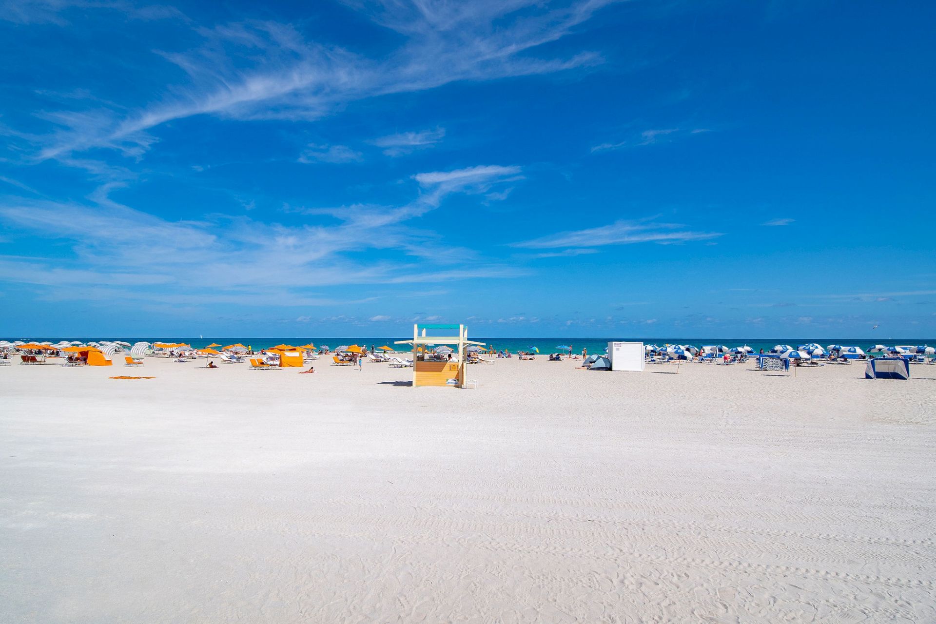A sandy beach with scattered umbrellas and chairs under a clear blue sky and distant ocean view.