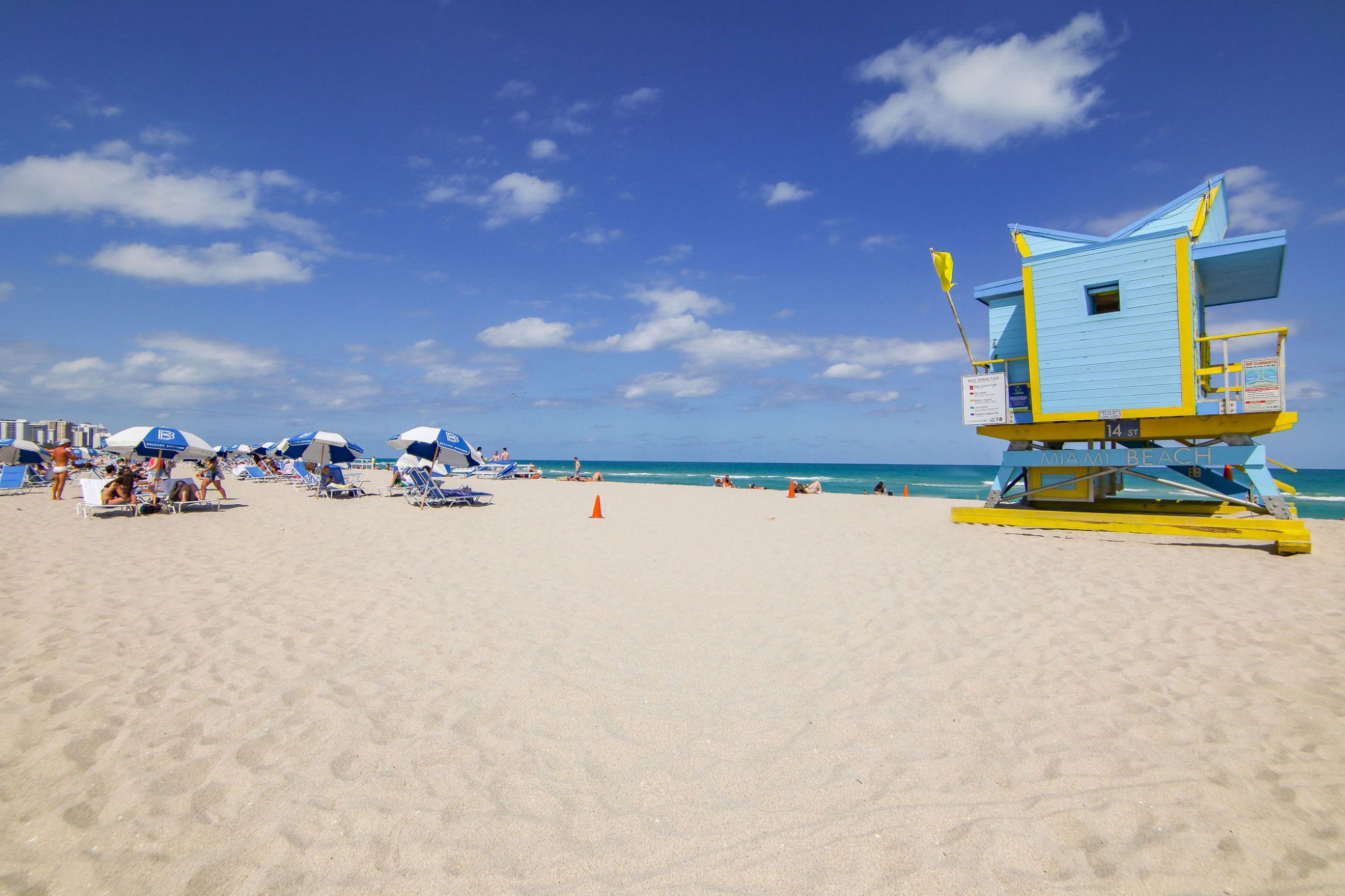 A sandy beach with umbrellas, lounge chairs, and a colorful lifeguard tower under a blue sky with scattered clouds by the ocean.