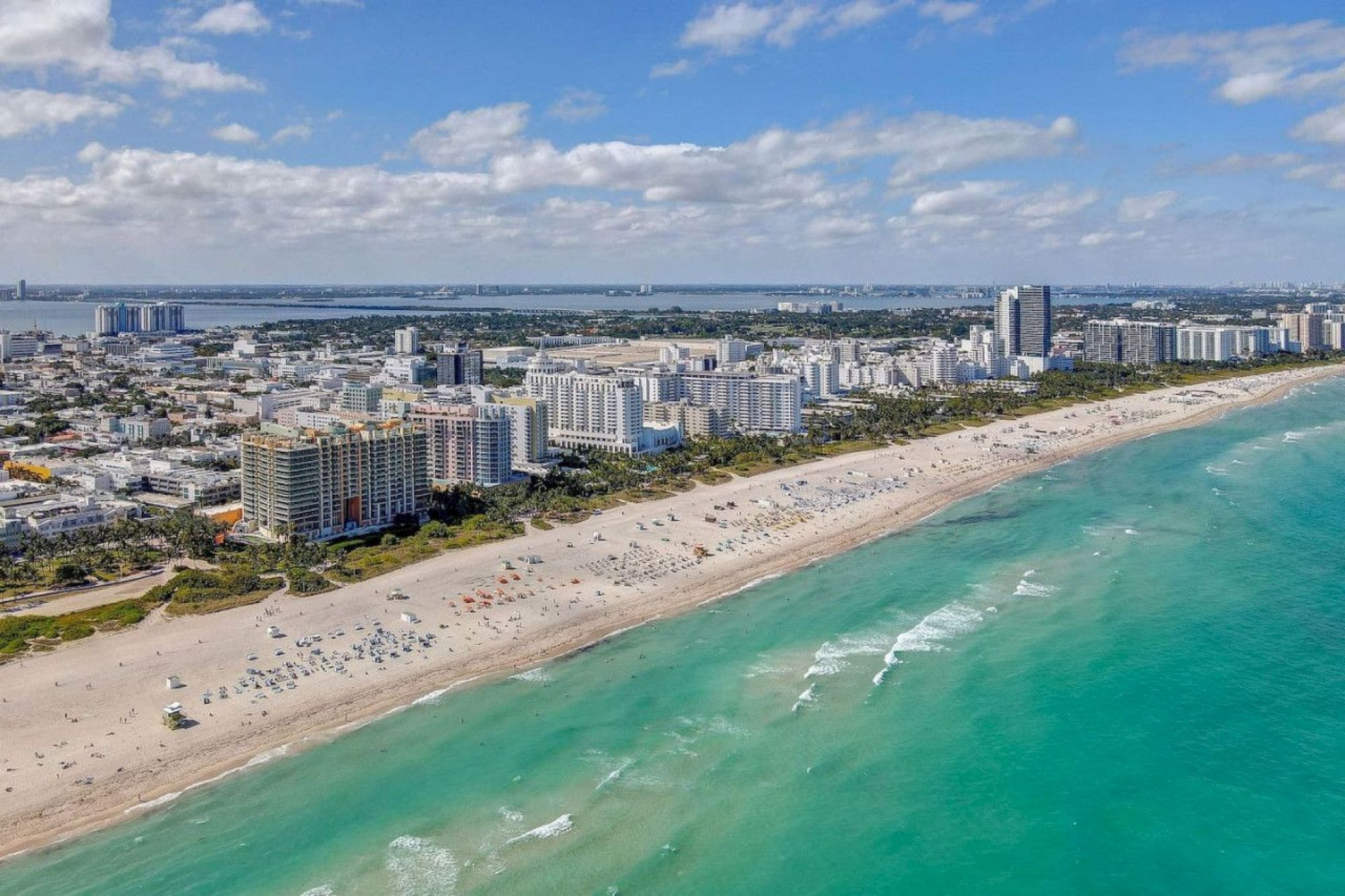 Aerial view of a coastal city with tall buildings, sandy beach, and turquoise ocean under a partly cloudy sky.