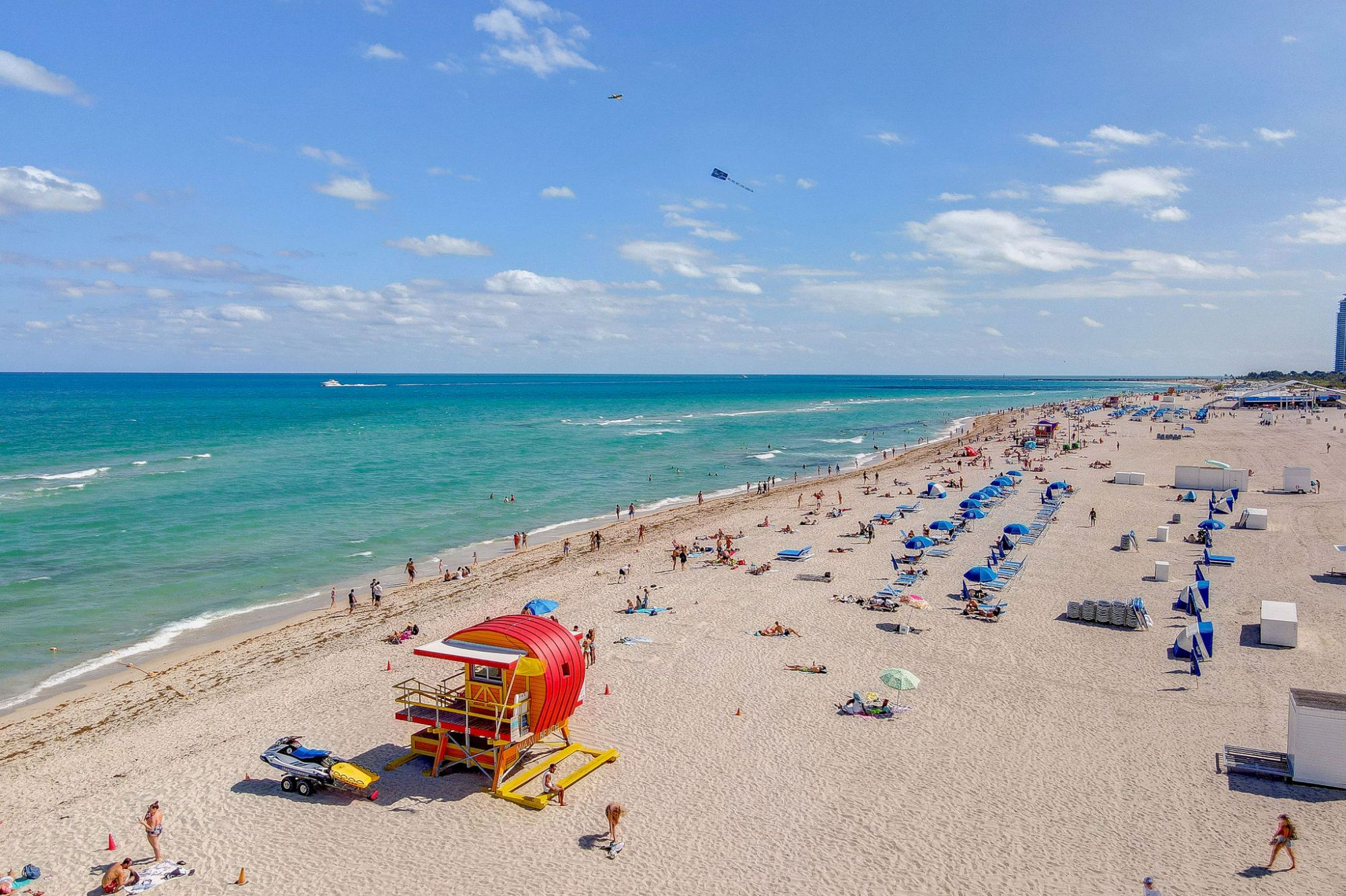 A sandy beach with people, a red lifeguard station, blue umbrellas, and the ocean under a partly cloudy sky.