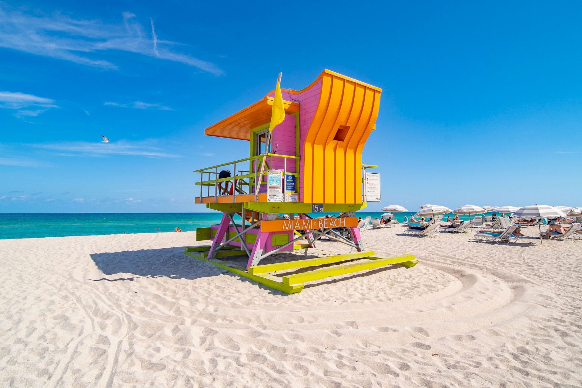 A colorful lifeguard tower stands on a sandy beach near the ocean, with sun loungers and umbrellas in the background under a clear blue sky.