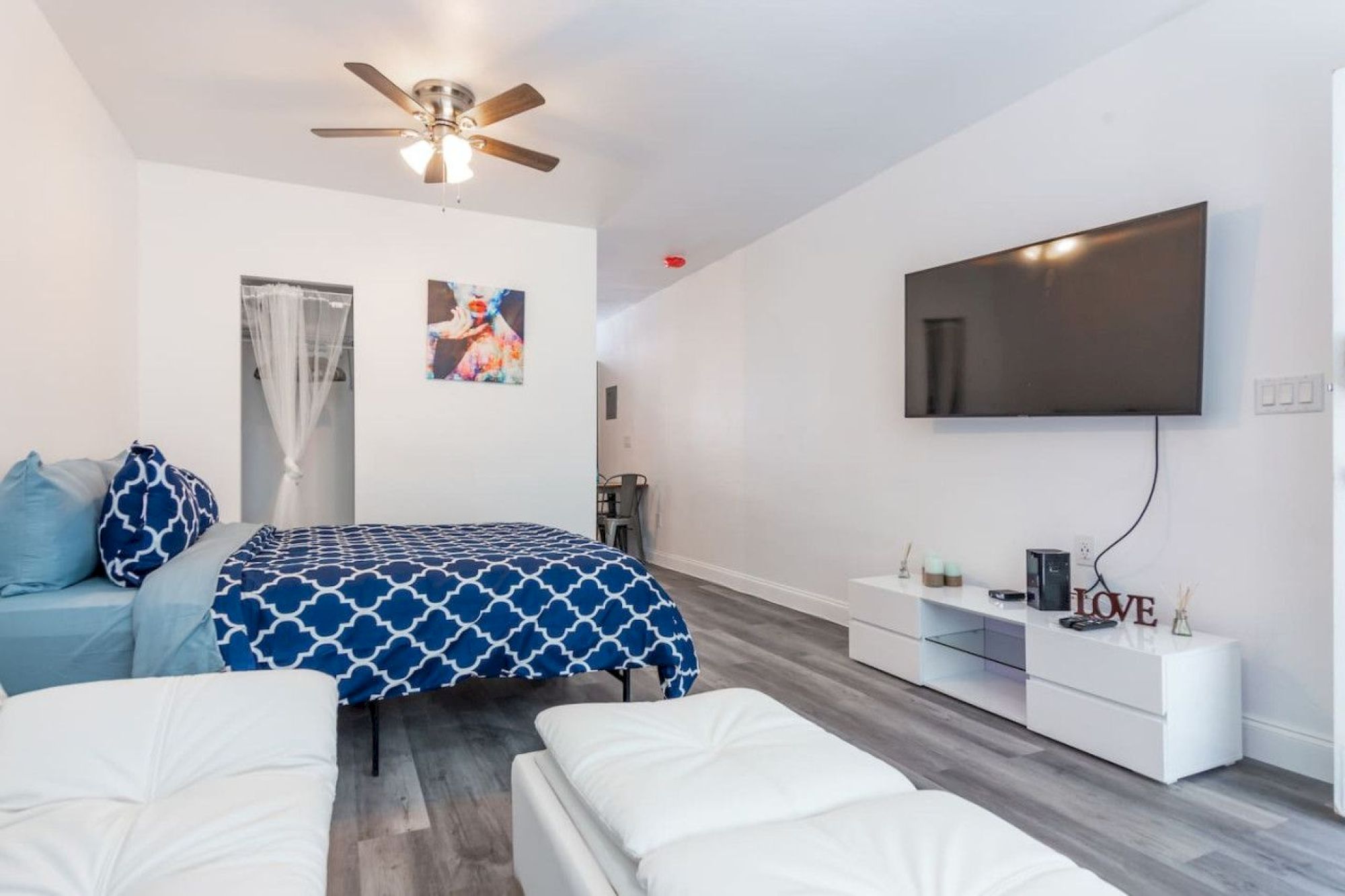 A modern room with a bed, TV, ceiling fan, and minimal decor. Blue accents on the bedspread and a small entertainment unit below the TV.