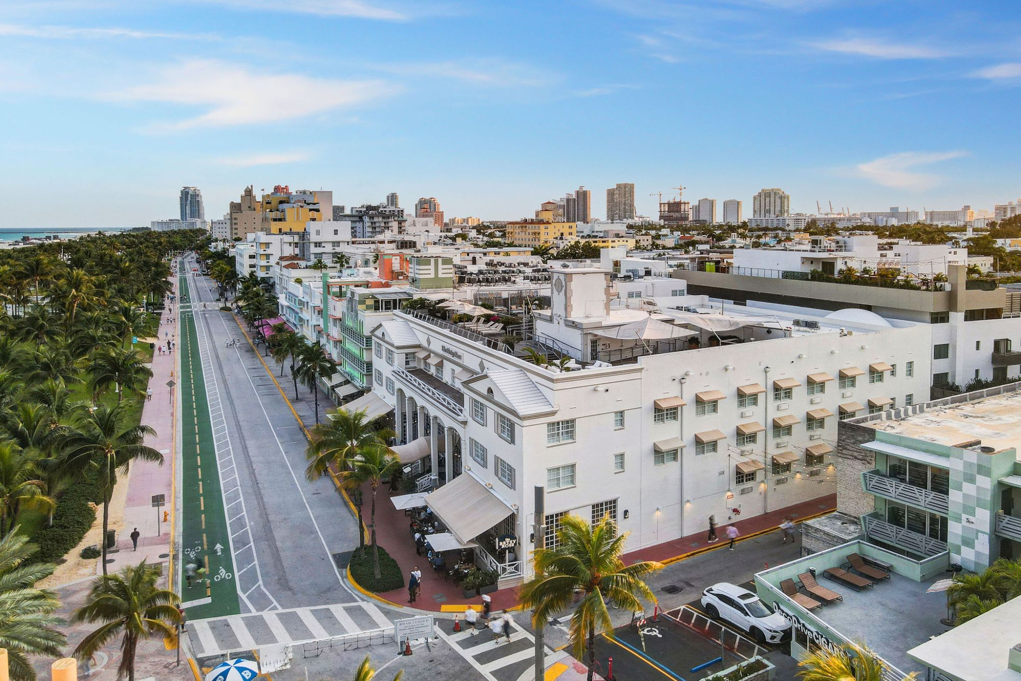 A coastal cityscape with a palm-lined road, white buildings, and a distant view of the ocean under a clear sky.