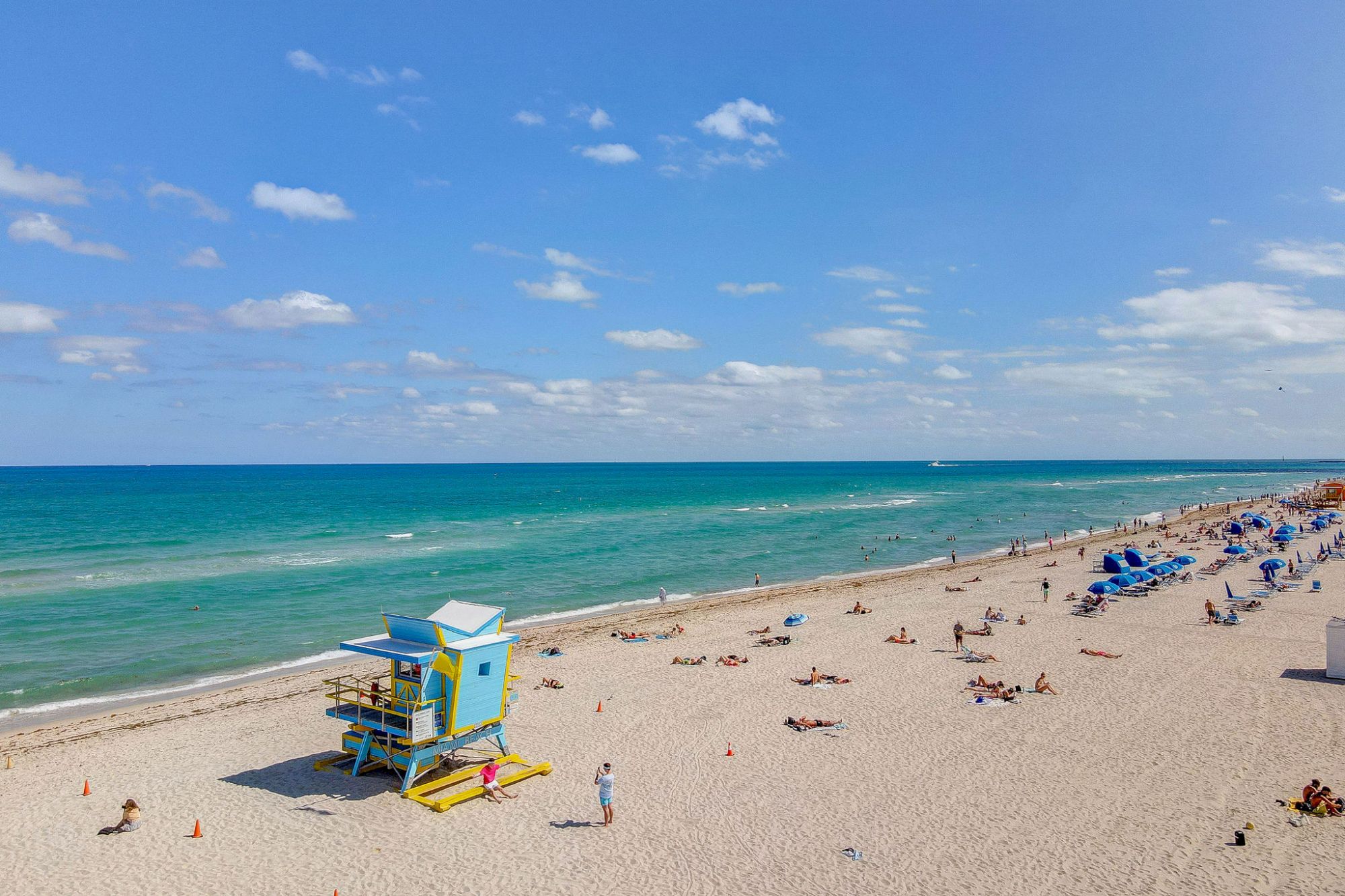A sandy beach with a blue lifeguard tower, people sunbathing, and a vibrant ocean under a clear blue sky.
