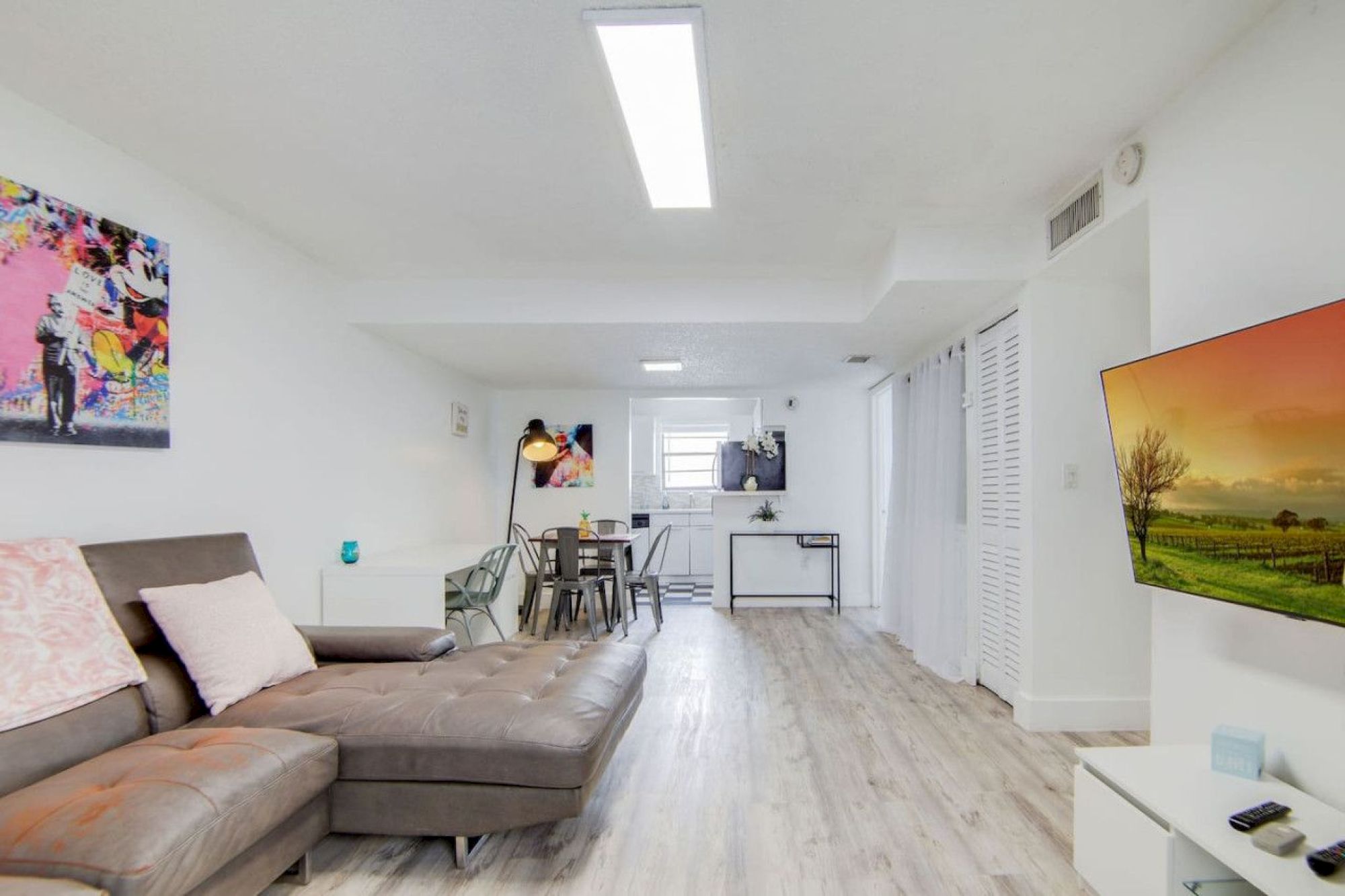 A bright living space with a sectional sofa, wall art, a dining area, and a TV. Modern decor and light wood flooring are featured.