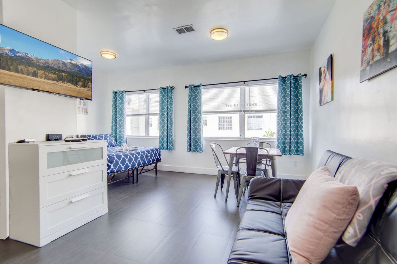 The image shows a bright living space with a bed, a sofa, a small dining table, blue curtains, a dresser, and a wall-mounted TV.