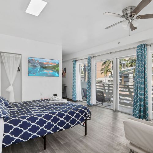 A bedroom with a bed, blue curtains, a ceiling fan, and large windows leading to a balcony, decorated with a coastal theme.