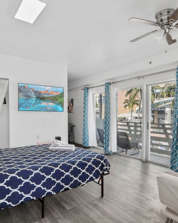 A bedroom with a bed, blue curtains, a ceiling fan, and large windows leading to a balcony, decorated with a coastal theme.