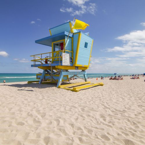 A colorful lifeguard tower on a sandy beach with people, ocean, and blue sky in the background.