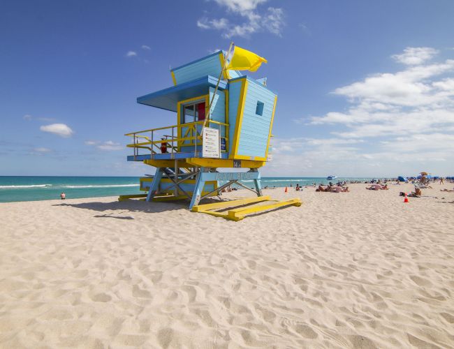 A colorful lifeguard tower on a sandy beach with people, ocean, and blue sky in the background.