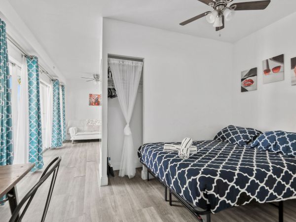 The image shows a modern bedroom with a bed, patterned bedding, wall art, curtains, and a ceiling fan. A small dining area is visible in the corner.