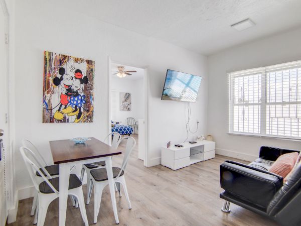 A bright living room with a dining table, wall art, mounted TV, and a black couch, featuring large windows allowing natural light.