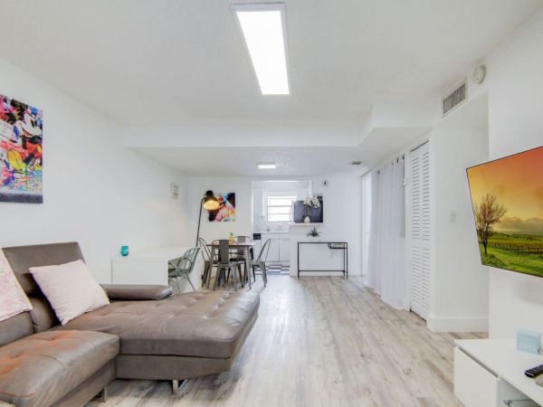 A modern living room with a gray couch, colorful art, a mounted TV, dining table, and white walls, offering a bright, open space feel.