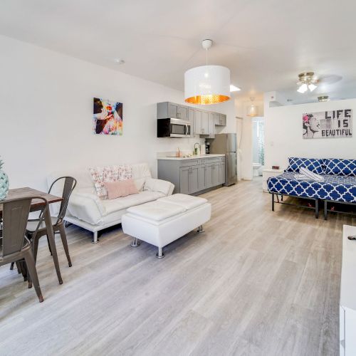 A modern living space with light wood flooring, kitchen, dining set, white and blue couches, artwork, and TV on the wall.