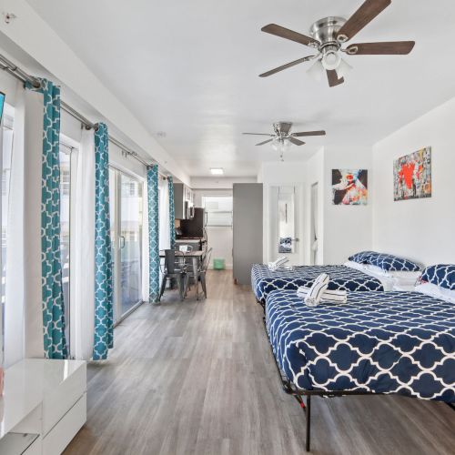 The image shows a bright, modern room with two beds, patterned bedding, ceiling fans, a TV, and a small dining area in the background.