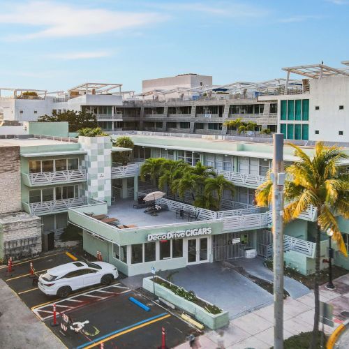 The image shows a modern building complex with palm trees, parked cars, and a clear sky.