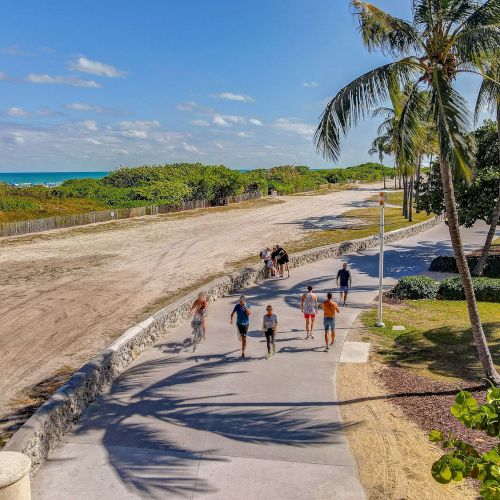 People are walking on a path near a sandy beach with palm trees and ocean in the background, under a partly cloudy blue sky.