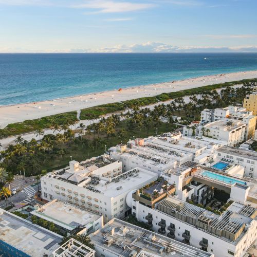 Aerial view of a beachfront cityscape with buildings, greenery, and a sandy beach beside a calm blue sea under a clear sky.