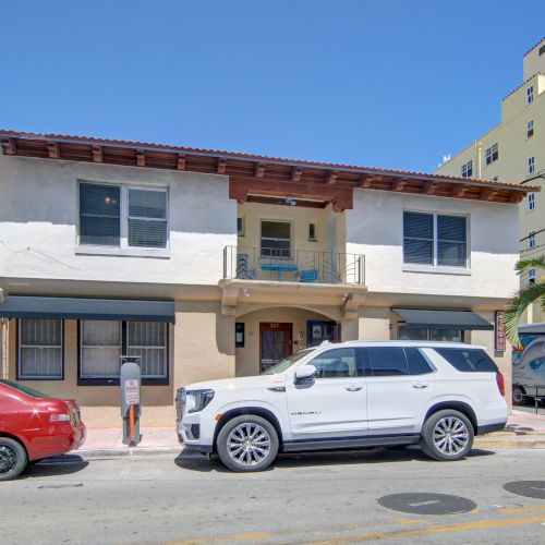 A two-story building with cars parked nearby, set against a clear blue sky, and a palm tree.