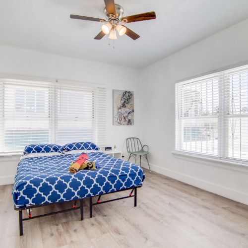 A bright bedroom with a blue-patterned bedspread, large windows, a ceiling fan, and minimal decor.
