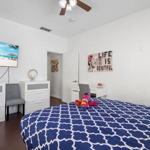 A bedroom with a blue-patterned bedspread, TV, dresser, and wall art. There are chairs, a desk, and decorations in a bright space.
