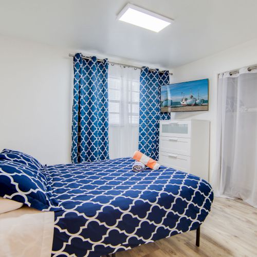 A bedroom with blue patterned bedding, matching curtains, a white dresser, wall art, and a TV. Light wooden floor and bright lighting.