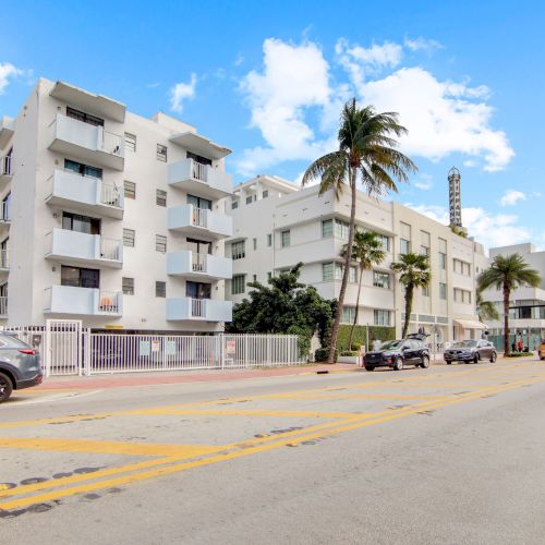 The image shows a street with modern buildings, palm trees, and parked cars under a clear blue sky with clouds.