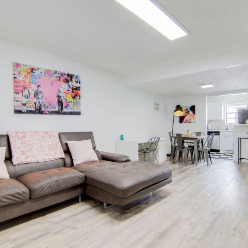 A modern living room with a brown sofa, colorful wall art, dining area, and TV on a stand on wooden flooring.