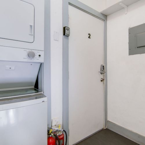 The image shows a white hallway with a door labeled "2," a wall-mounted washer-dryer unit, a fire extinguisher, and an electrical panel.