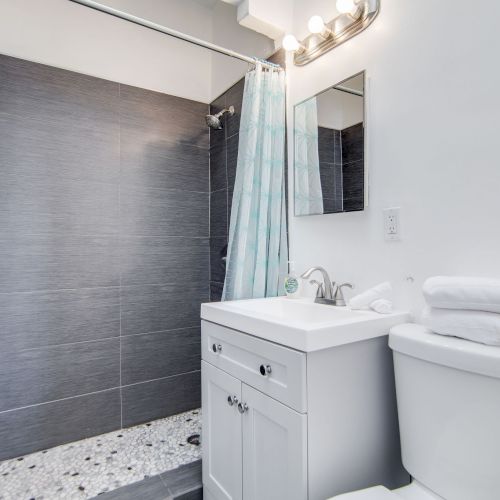 A small bathroom with a shower, sink, mirror, and toilet. Grey tiles in the shower, white fixtures, and folded towels on the toilet.