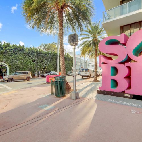The image shows a street scene with a large pink "SOBE" sculpture, palm trees, and nearby buildings in a sunny area.