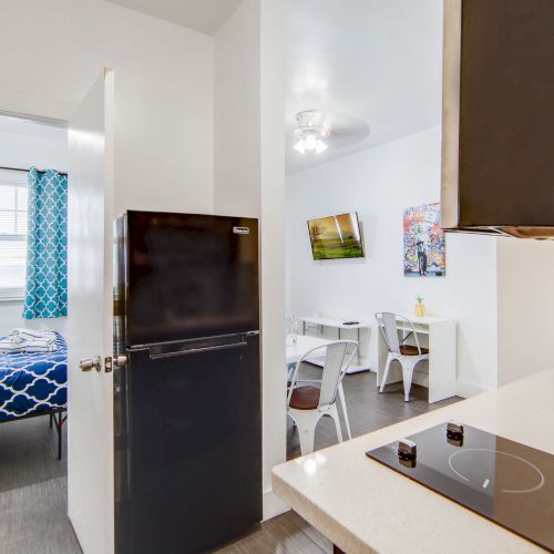 The image shows a compact apartment with a kitchen, a small dining area, and a bedroom with a blue-patterned bedspread and curtains.