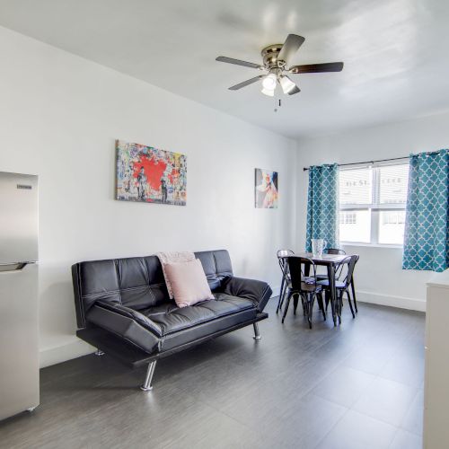 The image shows a modern living room with a sofa, dining table, fridge, wall art, ceiling fan, and a TV, all under bright light.