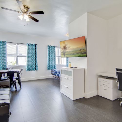 A bright living space with a sofa, desk, dining table, TV, and decorative curtains. The room has modern furnishings and a ceiling fan.