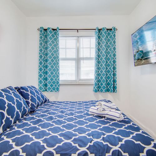 A cozy bedroom features a bed with blue patterned bedding, matching curtains, wall art, and a large window, creating a bright atmosphere.