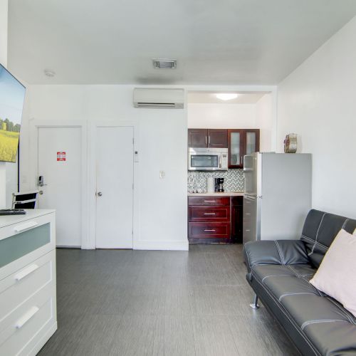 The image shows a modern living room with a wall-mounted TV, sofa, kitchenette, and vibrant artwork on the wall.