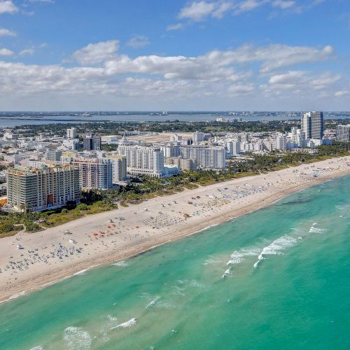 Aerial view of a coastal city with beachfront, high-rise buildings, and turquoise ocean under a partly cloudy sky.