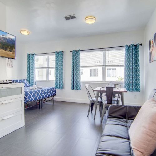 The image shows a modern room with a bed, TV, table, chairs, and couch, featuring blue curtains and wall art.