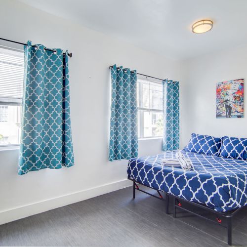 A bedroom with blue-patterned curtains and bedding, a desk with a chair, white walls, a window, and a nightstand next to the bed.