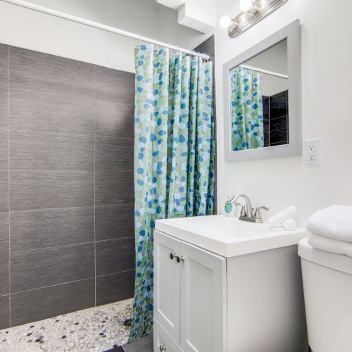 The image shows a bathroom with a shower, floral curtain, vanity with sink, mirror, and toilet with folded towels on top.