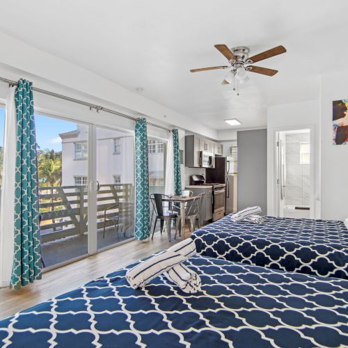 A bright room with two beds, a ceiling fan, blue curtains, balcony, and kitchenette, adorned with artwork, exuding a cozy, modern vibe.