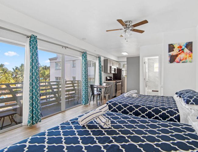 A bright room with two beds, a ceiling fan, blue curtains, balcony, and kitchenette, adorned with artwork, exuding a cozy, modern vibe.
