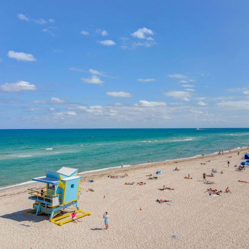 A beach scene with a blue lifeguard tower, people sunbathing, and umbrellas under a clear sky with scattered clouds.