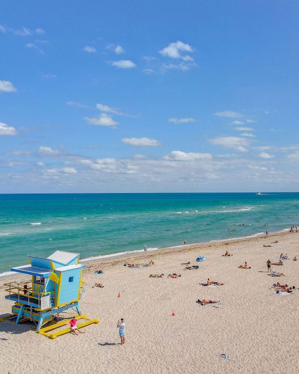 A beach scene with a blue lifeguard tower, people sunbathing, and umbrellas under a clear sky with scattered clouds.