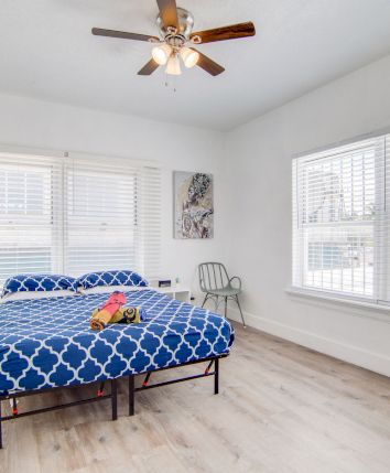 A bright bedroom with a blue-patterned bed, large windows, wooden floor, ceiling fan, and chairs, creating a cozy, inviting atmosphere.