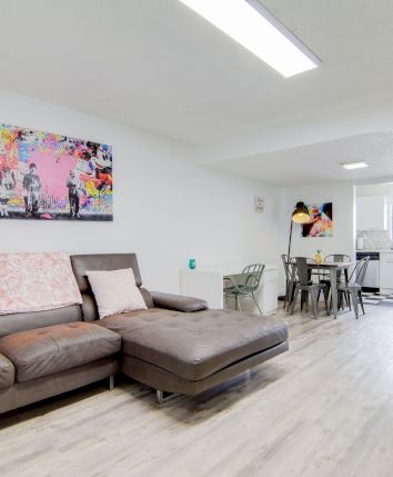 A modern living space with a brown sectional sofa, wall art, dining table, TV, and light wood flooring. Bright and minimalistic design.