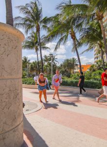 People are walking on a sunny path next to a large stone sign for Lummus Park in Miami Beach, surrounded by palm trees and buildings.