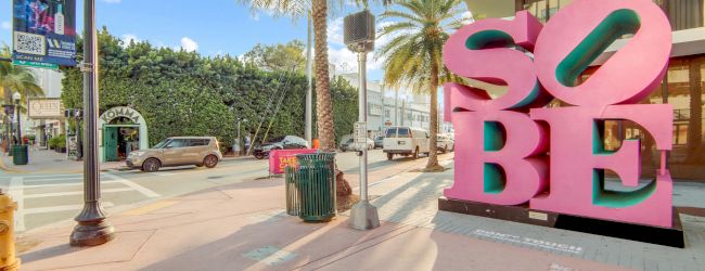 A street scene with palm trees, cars, buildings, and a large pink "SOBE" sculpture.