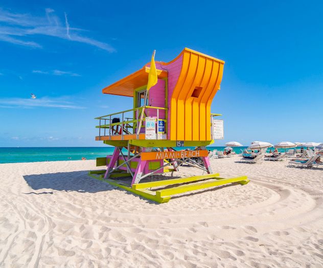A vibrant lifeguard tower in orange and purple sits on a sandy beach with the ocean and sun loungers in the background.