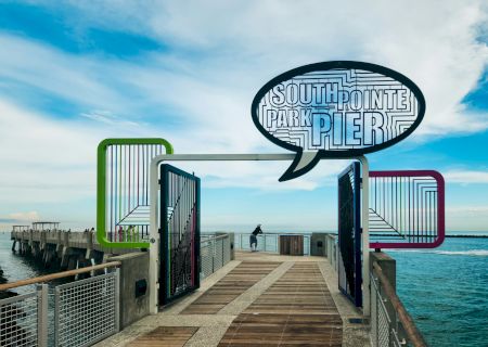 The image shows South Pointe Park Pier with a vibrant sign and clear sky. A person stands at the end of the pier, overlooking the water.